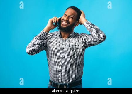 Enttäuschter Schwarzer, der einen Anruf hatte, der sich seinen Kopf schnappte, während er im Studio vor blauem Hintergrund stand Stockfoto