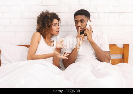 Ernster Schwarzer Mann, der Smartphone mit Zeigefinger auf den Lippen benutzt und schwarze Freundin mit lockigem Haar ignoriert, während er unter weißer Decke auf dem Bett um ... sitzt Stockfoto