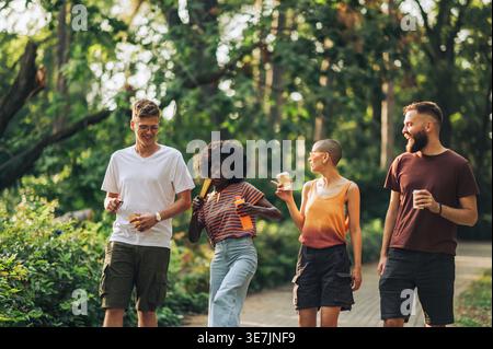 Schwarze Frau mit lockigem Afro, die eine Flasche Saft hält, während sie den Fächer als Mikrofon benutzt, und Spaß mit kaukasischen Freunden hat, die zusammen in Par... Stockfoto