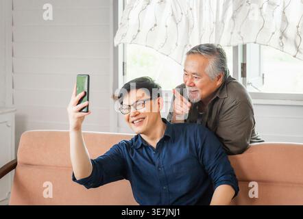 Asiatischer älterer Vater, der hinter einem Mann mittleren Alters steht und ein Smartphone benutzt, das auf der Couch sitzt, während beide im Wohnzimmer zu Hause Videoanrufe führen Stockfoto