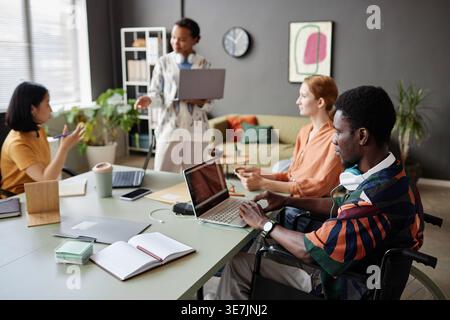 Afroamerikanischer Geschäftsmann im Rollstuhl mit Laptop bei Treffen mit verschiedenen Kollegen im Büro Stockfoto