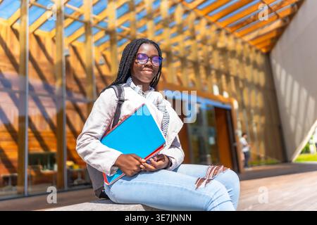 Schwarze Teenagerin mit Brille und Büchern, die tagsüber auf Beton vor dem Universitätscampus sitzen Stockfoto
