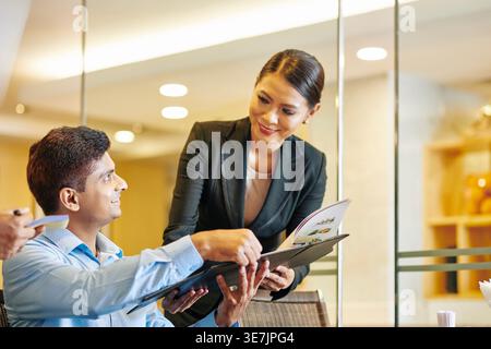 Asiatische Managerin nimmt Bestellungen von männlichen Kunden entgegen, die am Tisch im Hotelrestaurant sitzen Stockfoto