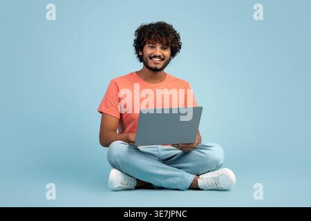 Lächelnder bärtiger indischer Freelancer mit lockigen Haaren, der auf dem Laptop vor blauem Hintergrund im Studio sitzt Stockfoto