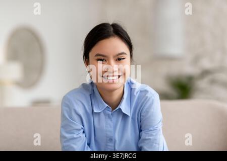 Lächelnde asiatische Frau in blauem Hemd, die zu Hause im Wohnzimmer auf der Couch sitzt Stockfoto