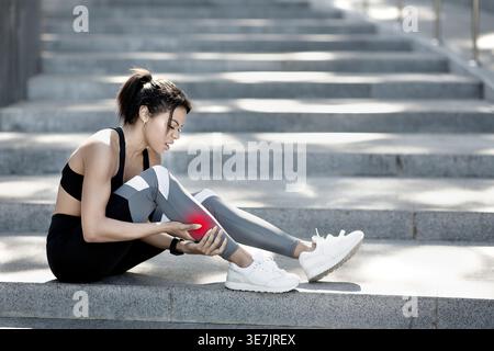 Schwarze Sportlerin in Sportkleidung massiert ihr verletztes Bein, während sie an sonnigen Tagen auf einer Treppe im Park sitzt Stockfoto