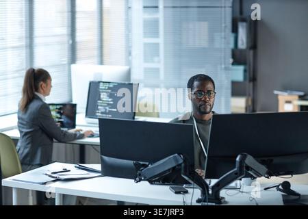 Schwarzer Programmierer, der eine Brille trägt und an Computern arbeitet, sitzt am Tisch neben einer kaukasischen Kollegin im Büro Stockfoto