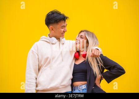 Lächelnde asiatische und kaukasische Paare mit Kopfhörern um den Hals, die sich umarmen und anschauen, während sie vor gelbem Hintergrund stehen... Stockfoto