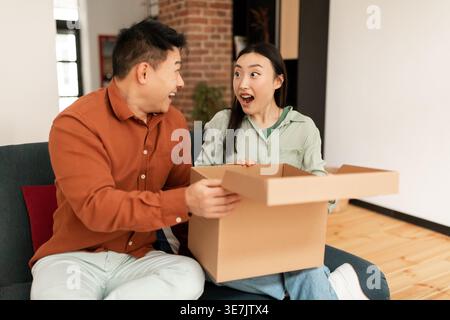 Glückliches asiatisches Paar öffnet die Lieferbox und sieht sich gegenseitig an, während es zu Hause auf dem Sofa sitzt Stockfoto