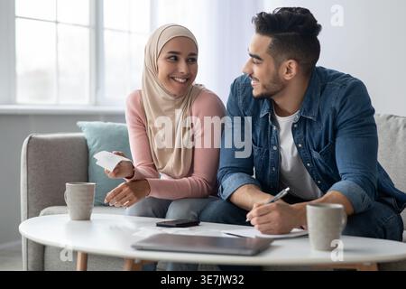 Lächelndes Paar aus dem Nahen Osten, das sich ansieht, wie man das Budget verwaltet, während man zu Hause mit Rechnungen am Tisch sitzt Stockfoto