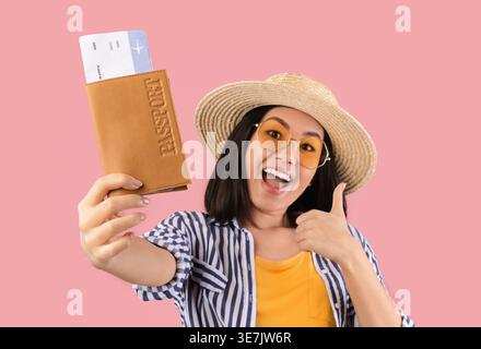 Fröhliche asiatische Frau, die Strohhut und Sonnenbrille trägt, einen Pass mit Flugtickets hält und die Daumen hoch zeigt, während sie gegen rosa Hintergras steht... Stockfoto