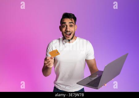 Aufgeregter arabischer Mann mit Bart, der Laptop und Kreditkarte hält, der vor rosa und violettem Farbverlauf im Studio steht Stockfoto