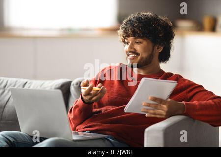 Asiatischer Mann mit Videoanruf auf Laptop mit Stift und Notizbuch, der zu Hause auf dem Sofa sitzt Stockfoto
