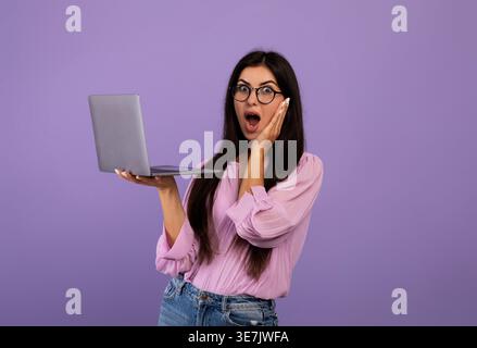Schockierte armenische Frau, die eine Brille trägt und einen Laptop hält, während sie im Studio vor violettem Hintergrund steht Stockfoto