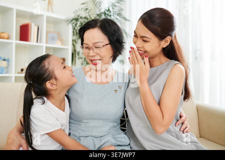 Ältere vietnamesische Großmutter in Brille umarmt ihre Tochter und Enkelin, während sie zusammen auf dem Sofa im Wohnzimmer zu Hause sitzt Stockfoto