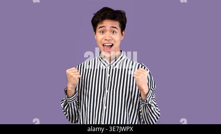 Asiatischer Mann mit gestreiften Polo-langen Ärmeln, der seine Fäuste zusammendrückt, während er im Studio mit offenem Mund gegen die violette Wand steht Stockfoto
