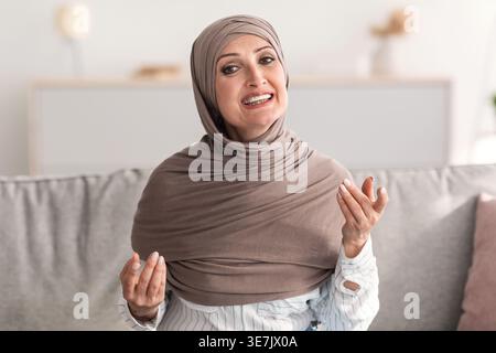 Eine muslimische Frau mittleren Alters im Hijab spricht und Gesten macht, während sie zu Hause auf dem Sofa sitzt Stockfoto