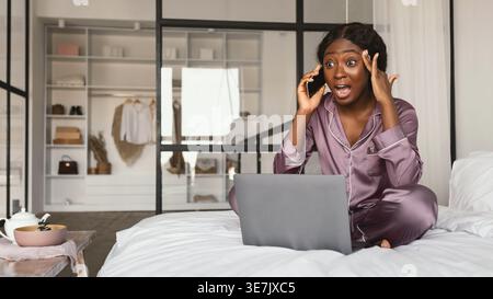 Schwarze Frau mit Seidenschlafanzug, die zu Hause im Schlafzimmer mit Laptop auf dem Bett sitzt Stockfoto