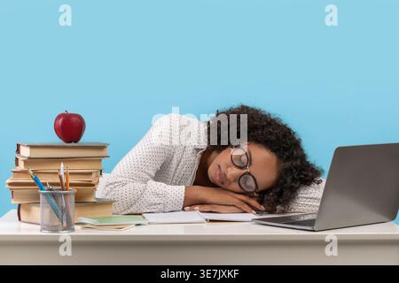 Müde Schwarze Teenager mit Brille und geschlossenen Augen sitzen und sich auf dem Tisch mit Laptop und Lernmaterialien vor blauem Hintergrund lehnen... Stockfoto