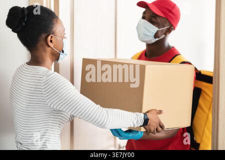 Schwarzer Liefermann mit Gesichtsmaske, der den Paketkasten an weibliche Kunden übergibt, während er während der Pandemie vor der Haustür zu Hause steht Stockfoto