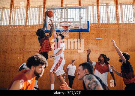 Schwarzer männlicher Baskteballspieler taucht den Ball in einen Basketballkorb ein, während der Teamkollege ihn während des Trainings auf dem Court blockiert Stockfoto