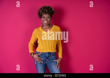Schwarze Frau mit lockigem Afro in gelbem Pullover und Jeans, die vor rotem Hintergrund im Studio steht Stockfoto