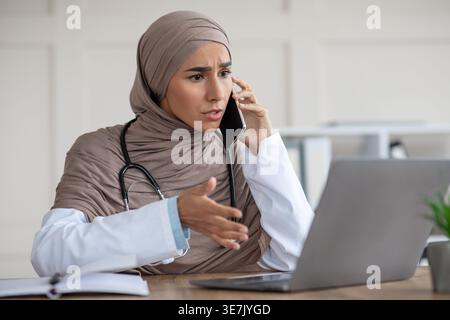 Nahöstliche Ärztin im Hijab mit Stethoskop am Hals, telefoniert mit Blick auf Laptop-Bildschirm am Tisch in der Klinik Stockfoto