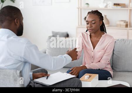 Schwarzer männlicher Psychologe hält ein Klemmbrett und gibt weiblichen Klienten ein Glas Wasser, während sie während einer privaten Therapiesitzung auf der Couch im Büro sitzen Stockfoto