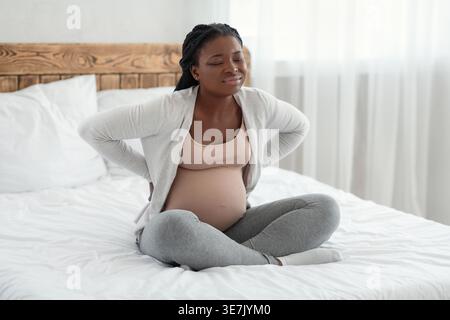 Schwarze schwangere Frau, die den Bauch berührt und unter Schmerzen leidet, während sie mit geschlossenen Augen auf dem Bett im Schlafzimmer zu Hause sitzt Stockfoto