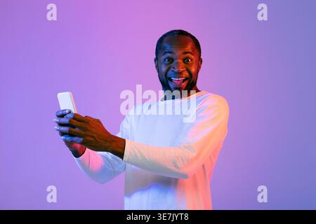 Lächelnder Schwarzer Mann mit Smartphone, während er vor violettem Hintergrund im Studio mit Neonlichtern steht Stockfoto