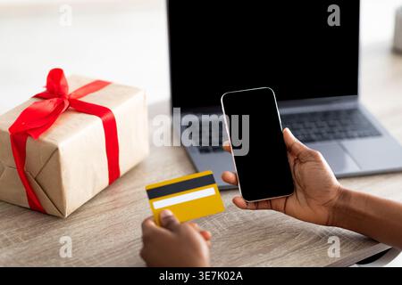 Zugeschnittenes Bild von Black Woman, die online mit Kreditkarte auf dem Smartphone bezahlt, während sie am Tisch mit Laptop und Paketfach im Wohnzimmer zu Hause sitzt Stockfoto