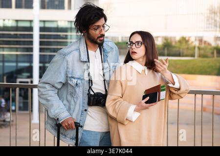 Asiatisches und kaukasisches Paar mit Brille, die Reisedokumente hält, während es dicht neben dem Gepäck vor dem Flughafen steht Stockfoto