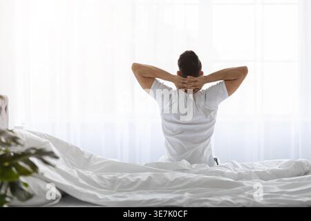 Ein arabischer Mann sitzt mit Händen hinter dem Kopf auf dem Bett im Schlafzimmer zu Hause Stockfoto