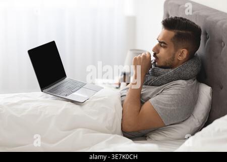 Arabischer Mann hustet in der Hand und benutzt Laptop, während er zu Hause auf dem Bett sitzt Stockfoto