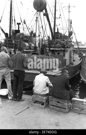 Krim, Gurzuf oder Jalta, 1968, Seehafen. Drei Männer entspannen sich auf dem geschäftigen Betonpier, zwei von ihnen sitzen bequem auf Holzkisten, während sie rauchen und plaudern. Sie befinden sich direkt neben stabilen, vertäuten Fischerbooten, die mit komplexen Takelagen, Netzen und schwerer nautischer Ausrüstung ausgestattet sind. Dieses sehr authentische Vintage-Archivfoto fängt die ruhige, hart arbeitende Atmosphäre der lokalen maritimen Industrie, typische Hafenaktivitäten am Meer und das friedliche tägliche Leben vor dem Krieg ein. Stockfoto