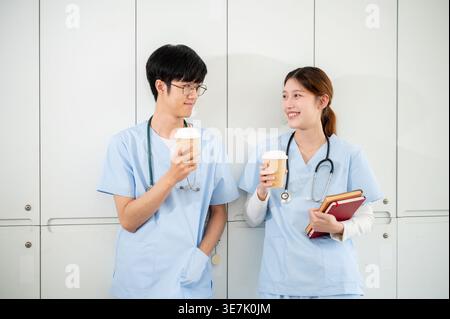 Asiatische männliche und weibliche Medizinstudenten, die Kaffee trinken und sich gegenseitig ansehen, während sie in der Umkleidekabine stehen Stockfoto