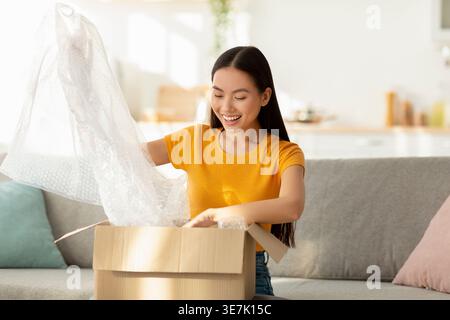 Asiatische Frau, die Papppaket auspackt, zufrieden mit Einkaufen und großem Einkauf, auf Sofa im Wohnzimmer sitzend Stockfoto