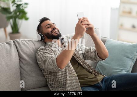 Arabischer Mann mit Bart, der Kopfhörer trägt und Musik mit dem Smartphone hört, während er sich im Wohnzimmer zu Hause auf dem Sofa zurücklehnt Stockfoto