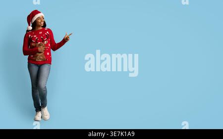 Lächelnde schwarze Frau mit Weihnachtsmütze und Pullover, die wegblickt und zeigt, während sie im Studio vor blauem Hintergrund steht Stockfoto