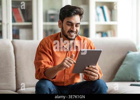 Positiver cooler, stilvoller junger Mann aus dem Nahen Osten, der im Wohnzimmer auf der Couch sitzt, zu Hause ein digitales Tablet verwendet und lächelt, mit Freunden chatet, Websu Stockfoto