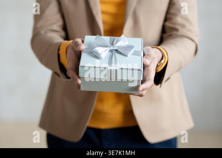 Zugeschnittenes Bild eines Mannes mit gemischter Rasse, der einen Mantel trägt und eine Geschenkbox hält, die im Studio vor grauem Hintergrund steht Stockfoto