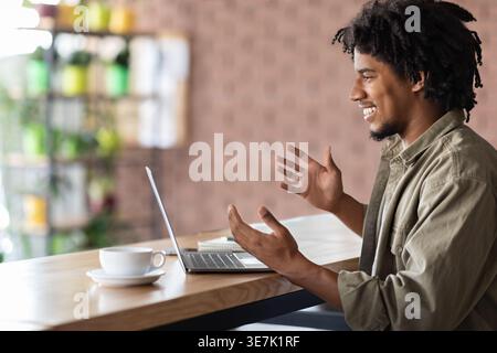 Lächelnder Schwarzer Mann mit afro-Gesten Händen, die Videoanrufe mit einem Laptop machen, der am Tisch im Café sitzt Stockfoto