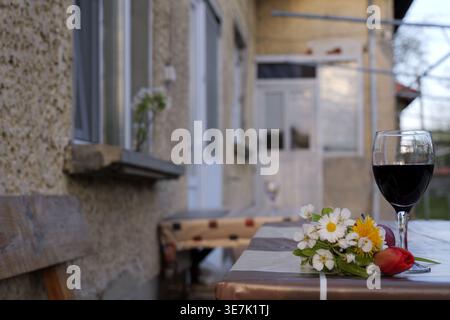 Ein Glas Rotwein mit Apfel und Wildblumen auf einer karierten Tischdecke, beleuchtet von warmem, goldenem Stundenlicht in einer ruhigen Umgebung im Freien. 2026 Stockfoto