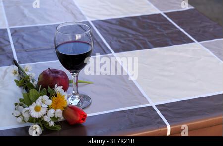 Stillleben mit einem Glas Rotwein, einem roten Apfel und einem Blumenstrauß auf einer karierten Tischdecke, draußen in der Nähe eines Weinbergs. 2026 Stockfoto