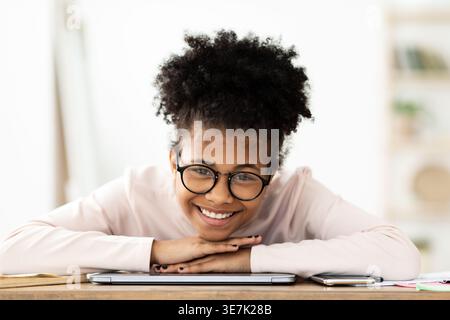 Lächelndes schwarzes Mädchen, das eine Brille trägt, das Kinn an den Händen über dem Laptop hängt und zu Hause am Tisch sitzt Stockfoto