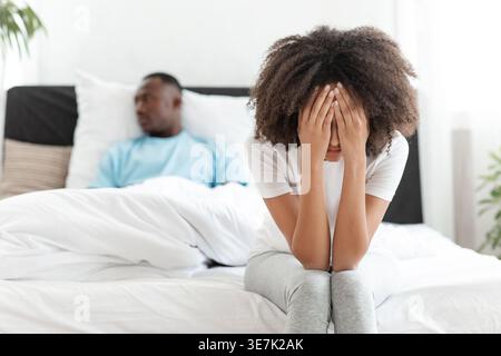 Schwarze Frau mit lockigen Haaren, die ihr Gesicht bedecken, während sie im Schlafzimmer zu Hause auf dem Bett gegen ihren Freund sitzt Stockfoto