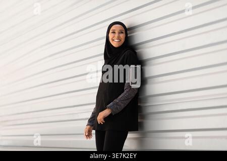 Lächelnde Athletin aus dem Nahen Osten, die tagsüber auf der Straße steht und einen schwarzen Hijab trägt Stockfoto