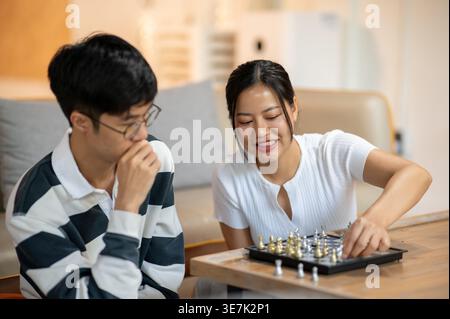 Asiatische Frau in weißem Hemd spielt Schach mit männlichem Freund auf Tisch im Wohnzimmer zu Hause Stockfoto