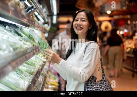 Thailändische Frau mit Schultertasche, die Gemüse aus dem Regal wählt, während sie im Supermarkt steht Stockfoto