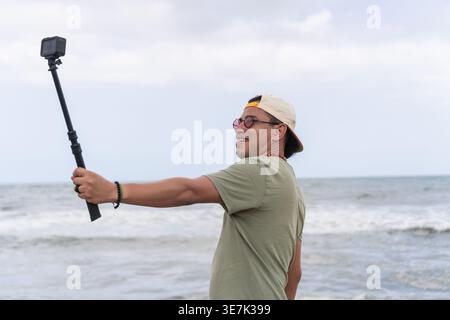 Hispanischer Mann mit Mütze und Sonnenbrille, der tagsüber Selfie vor der Kamera macht, während er am Strand steht Stockfoto